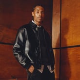 Un homme portant une veste en cuir noir de style bomber, posant devant un fond en bois sombre.