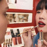 Une maquilleuse applique du rouge à lèvres au pinceau sur une jeune femme asiatique à frange.