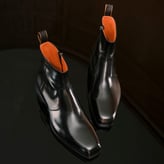 Une paire de bottines élégantes en cuir noir brillant avec une doublure intérieure orange vif est posée sur une surface sombre et réfléchissante.