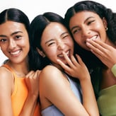 Trois jeunes femmes sourient et rient ensemble sur fond blanc. Elles portent des débardeurs colorés (orange, lilas, vert). La femme au centre plisse les yeux et porte ses doigts à sa bouche dans une expression de complicité joyeuse.