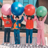 Quatre enfants debout sur un banc en béton, portant des tenues colorées et brandissant au-dessus de leur tête de gros ballons de fitness (rose, bleu, rouge, vert), avec un bâtiment moderne en arrière-plan.