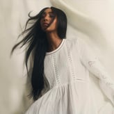 Une femme aux longs cheveux noirs balayés par le vent pose de profil. Elle porte une robe blanche plissée à détails brodés, sur un fond de tissu drapé clair.