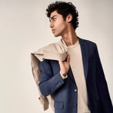 Un jeune homme aux cheveux bouclés pose de profil sur fond beige. Il porte un costume bleu sur un haut écru et maintient une veste beige claire sur son épaule droite.