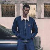 Un jeune homme à l'expression neutre, vêtu d'un ensemble veste et pantalon en denim bleu, pose les mains dans les poches devant une voiture.