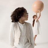 Un garçon aux cheveux bouclés regarde vers une fillette en robe blanche qui lève un grand ballon beige. Ils portent des vêtements clairs sur un fond épuré.