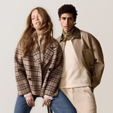 Un couple pose sur fond clair. La femme porte une veste à carreaux sur un jean bleu. L'homme, aux cheveux bouclés, porte une veste beige, un pull écru et un bandana autour du cou.