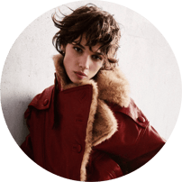 Portrait en contre-plongée d'une femme aux cheveux courts et ondulés, vêtue d'un manteau bordeaux en cuir à col en fourrure.
