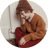 Enfant aux cheveux bouclés portant un bonnet tricoté, un gilet marron à boutons et un pantalon en velours cotelé terracotta.