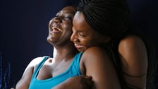 Deux femmes noires partagent un moment de complicité et de rire. L'une, en débardeur turquoise, rit aux éclats tandis que l'autre, coiffée de tresses, l'enlace par-derrière sur fond sombre.