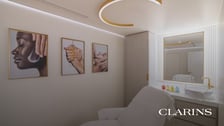 Cabinet de soin esthétique Clarins moderne et épuré, comprenant une table de massage blanche, un miroir avec lavabo et des cadres photo représentant des soins de la peau accrochés au mur.