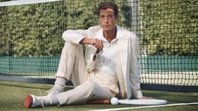 Un homme habillé de façon élégante est assis par terre sur un court de tennis extérieur, adossé contre le filet