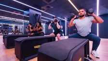 Quatre sportifs effectuent des squats synchronisés avec des haltères aux épaules dans une salle de fitness moderne. Ils se tiennent devant des bancs Technogym sous un éclairage de néons violets.