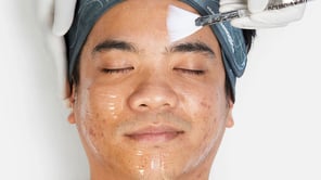 Gros plan sur le visage d'un homme aux yeux clos recevant un soin. Une personne gantée applique un produit translucide sur son front avec un pinceau plat blanc. L'homme porte un bandeau gris « Dermalogica ». Sa peau présente des imperfections.