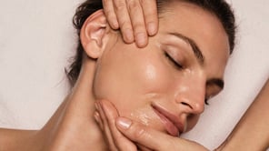 Gros plan sur une femme aux yeux clos, deux mains massant son visage enduit d'huile.