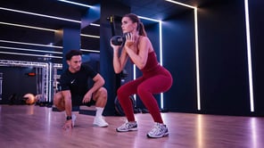 Une femme en ensemble de sport bordeaux effectue un squat avec un haltère dans une salle de fitness moderne. À ses côtés, un homme en tenue de sport noire l'observe en position accroupie. Le studio est décoré de lignes de néons blancs verticaux.