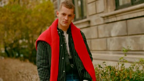 Un homme portant un pull rouge sur ses épaules, par-dessus une chemise rayée et une veste à rayures.