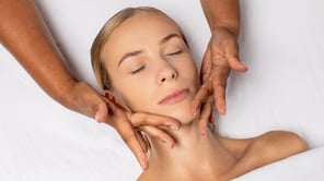 Vue aérienne d'une femme blonde aux yeux fermés, allongée et recevant un massage facial par une personne aux mains plus foncées.