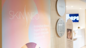 Vue en perspective de l'entrée d'un institut Skinneo. Au premier plan, une affiche aux tons pastels présente le logo et les expertises. Au mur, des panneaux circulaires affichent les tarifs. En arrière-plan, un écran bleu indique « CORPS ».