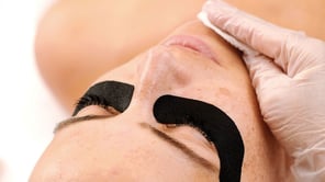 Gros plan sur le visage d'une personne aux yeux clos portant des patchs noirs sous les yeux. Une main gantée utilise une lingette blanche pour nettoyer le menton.