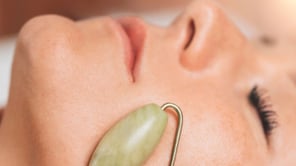 Gros plan sur le bas du visage d'une personne aux yeux clos, recevant un soin. Un rouleau de massage en pierre verte (jade) est appliqué sur sa joue. La peau présente de légères taches de rousseur et des cils noirs recourbés sont visibles.