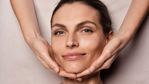 Gros plan frontal d'une femme souriante, le visage cadré par deux mains effectuant un soin.