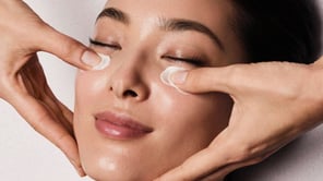 Gros plan sur une femme aux yeux fermés, recevant l'application d'une crème sur le contour des yeux.