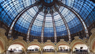Vue en contre-plongée de la majestueuse coupole en verre et fer forgé des Galeries Lafayette, surmontant des balcons ornés et des arches richement décorées de style Art nouveau.