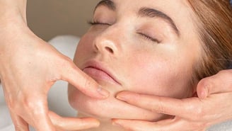 Gros plan sur une personne aux yeux clos recevant un massage du menton et de la mâchoire. Deux mains manipulent délicatement la peau du bas du visage. Le cadrage est serré sur le nez, la bouche et le menton.