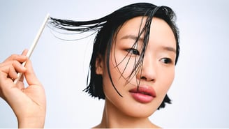 Gros plan sur une femme aux cheveux courts et mouillés, en train de démêler une mèche avec un peigne blanc sur un fond clair et épuré.