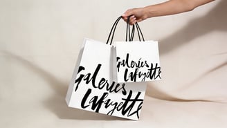 Un bras tendu tient deux sacs de shopping blancs à anses noires de tailles différentes, marqués d'une inscription noire « Galeries Lafayette », sur un fond beige drapé.