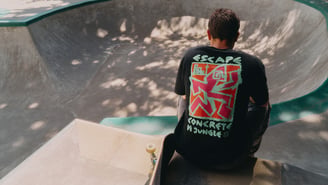 Vu de dos, une personne assise au bord d'un skatepark en béton porte un t-shirt noir avec un graphique coloré "Escape Concrete Jungle", son skateboard posé à côté de lui.