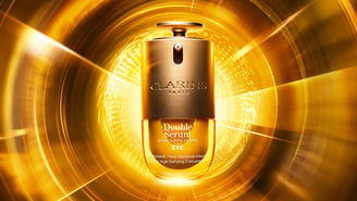 Un flacon du soin « Double Serum Eye » de Clarins Paris au centre, sur un fond doré lumineux et étincelant formant des cercles concentriques avec des faisceaux de lumière.