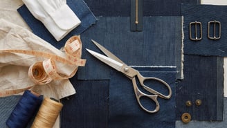 Vue aérienne d'une paire de ciseaux posée sur des morceaux de tissu en jean. On aperçoit aussi un mètre ruban, des bobines de fil, une fermeture éclair, des boutons et des boucles en métal.