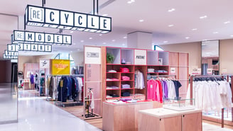 L'intérieur d'un magasin de vêtements lumineux avec des portants, des meubles en bois et de grandes enseignes lumineuses suspendues affichant les mots « RECYCLE », « RE[MODEL] » et « START ».