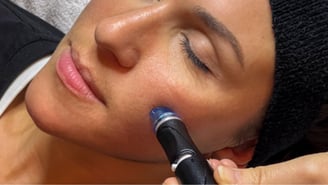 Une femme portant un bandeau noir reçoit un soin du visage. Une main manipule un stylet d'aspiration à embout bleu directement sur sa joue. Ses yeux sont fermés et elle repose sur une serviette blanche.