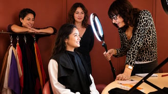 Dans un atelier de colorimétrie, une femme assise avec une cape noire sourit pendant qu'une conseillère en image ajuste une lampe anneau lumineuse (ring light) devant son visage.