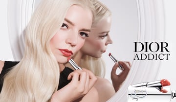 Une femme aux longs cheveux blond platine tient un tube de rouge à lèvres argenté devant un miroir rond. Son reflet apparaît de profil. À droite, l'inscription noire « DIOR ADDICT » surmonte un gros plan du produit de maquillage.