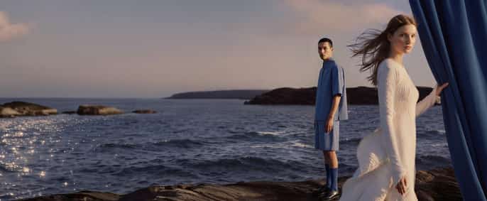 Une femme en robe blanche longue et un homme en ensemble bleu posent sur des rochers au bord de la mer au coucher du soleil, avec un grand rideau bleu sur la droite.