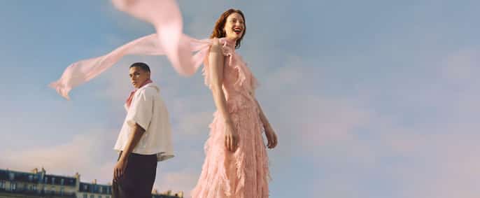 Une femme en robe rose frangée dont le foulard flotte au vent et un homme en chemise blanche posent en extérieur sous un ciel clair.