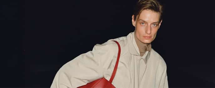 Un mannequin au regard fixe porte un manteau beige et un grand sac rouge vif qui tranchent sur un fond noir uni.