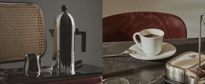 Une cafetière italienne en inox brillant et un petit pot à lait assorti sont posés sur un plateau métallique. L'ensemble est placé devant le dossier en cannage d'une chaise en bois. L'ambiance est feutrée avec des tons gris et naturels.